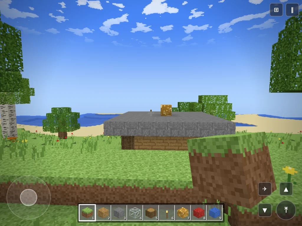 YoursCraft の画面。草原に板材と石レンガで建てた小屋が立ち、奥には砂浜と海、空にはドット絵の雲が浮かんでいる。下部にブロックを選ぶホットバー、左下に移動スティック、右下にジャンプと飛行のボタンがある